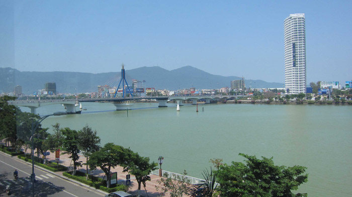 晴天の青空に、緑がかった色のハン川に架かるハン川橋（長さ400メートル程）の全景。遠くに山々が見え、橋の向こう側に37階建ての白い高層ホテル